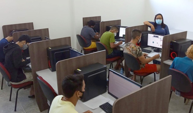 uma sala de informática, com várias pessoas usando computadores e uma instrutora, todos estão de máscara